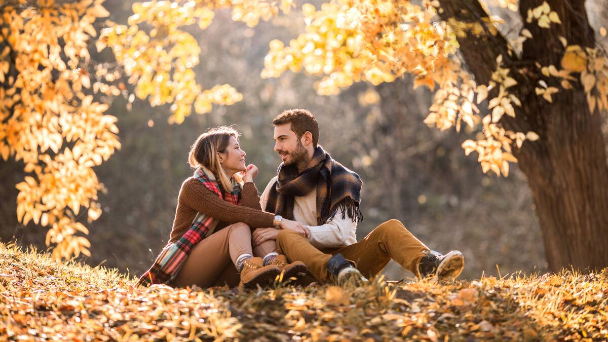 A couple sitting side by side outside in the autumn leaves, looking at each other happily.