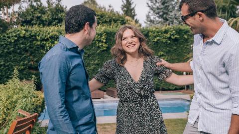 A woman standing with her two male friends, an arm out to each of them, all smiling as they chat.