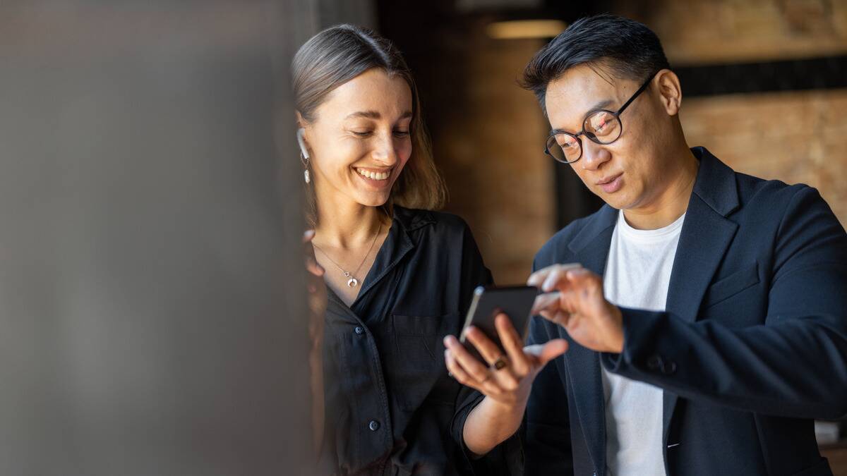 Two friends, a man and a woman, standing inside as the man shows the woman something on his phone.