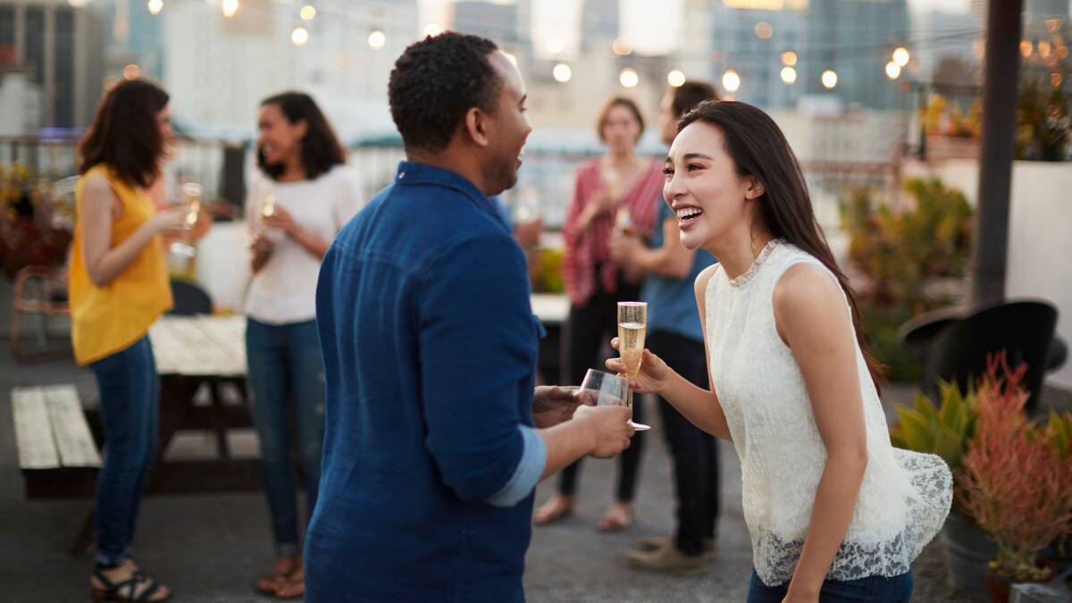 Two friends, a man and a woman, talking at a rooftop party, both holding drinks.