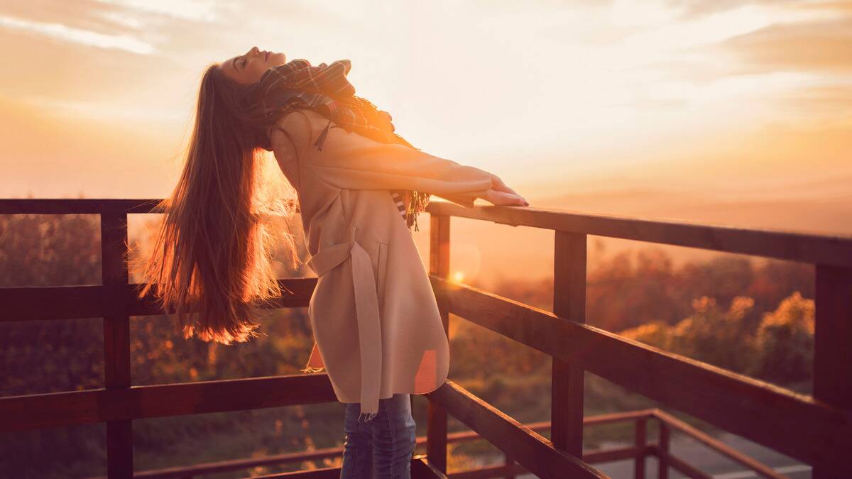 A woman holding onto a balcony rail and leaning back, smiling up at the sky, as the sun sets behind her.