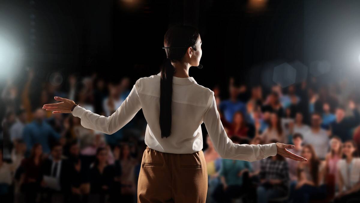 A woman, photographed from behind, on stage giving a presentation to a crowd.