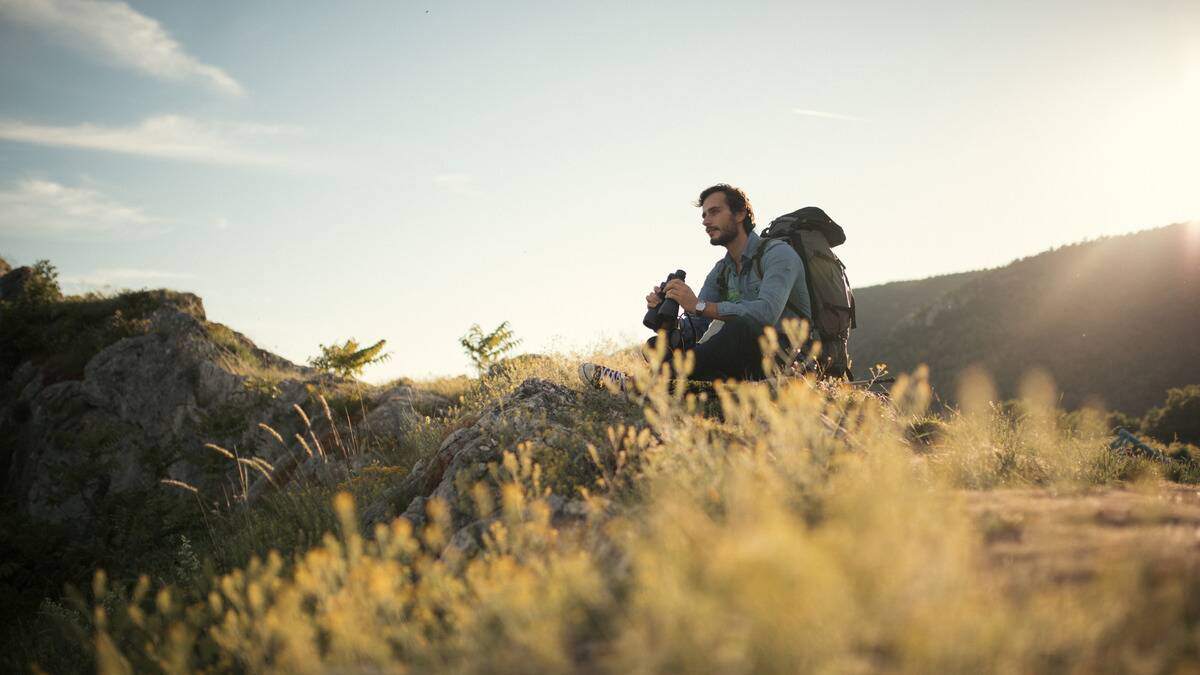  A man sitting outside on a hill range, holding a large pair of binoculars.