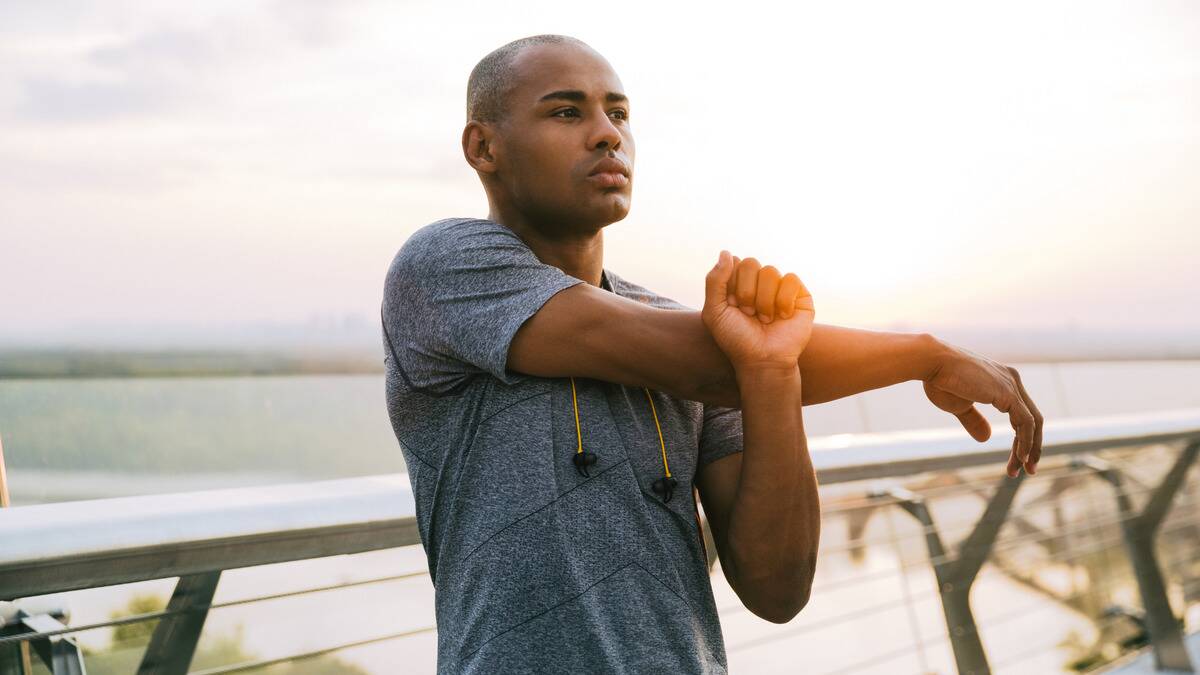 A man standing outside by a railing in front of he water, stretching his shoulders before he starts working out.