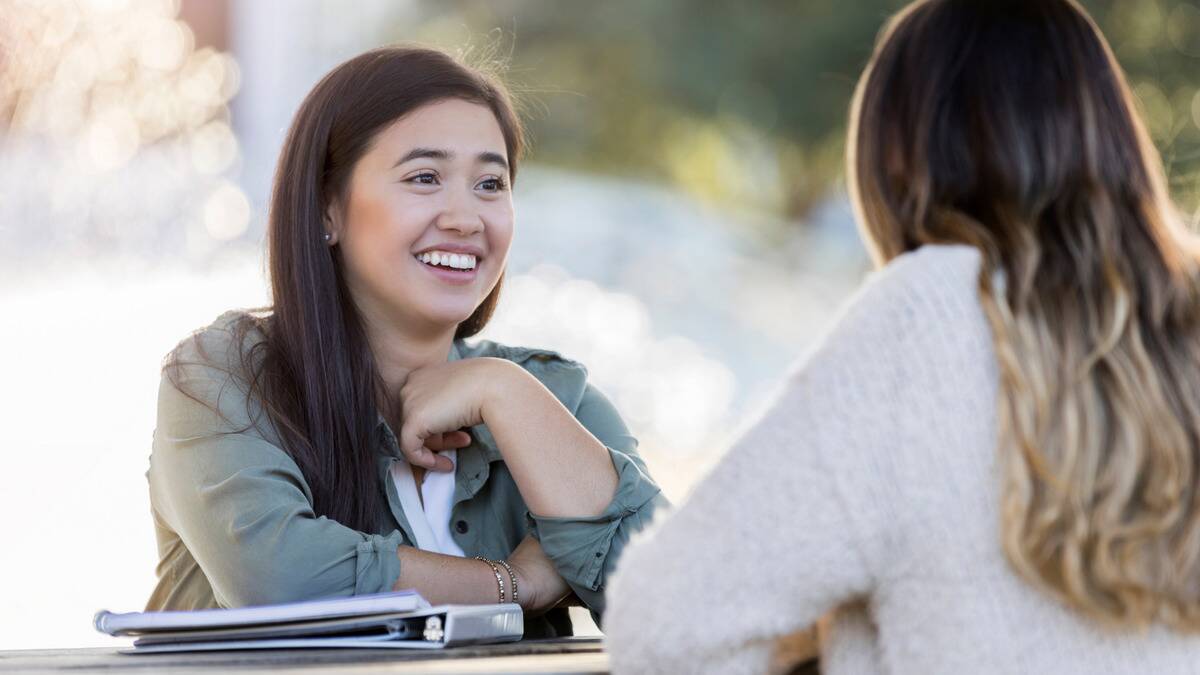 A woman sat across a table from her friend who she's speaking to while smiling.