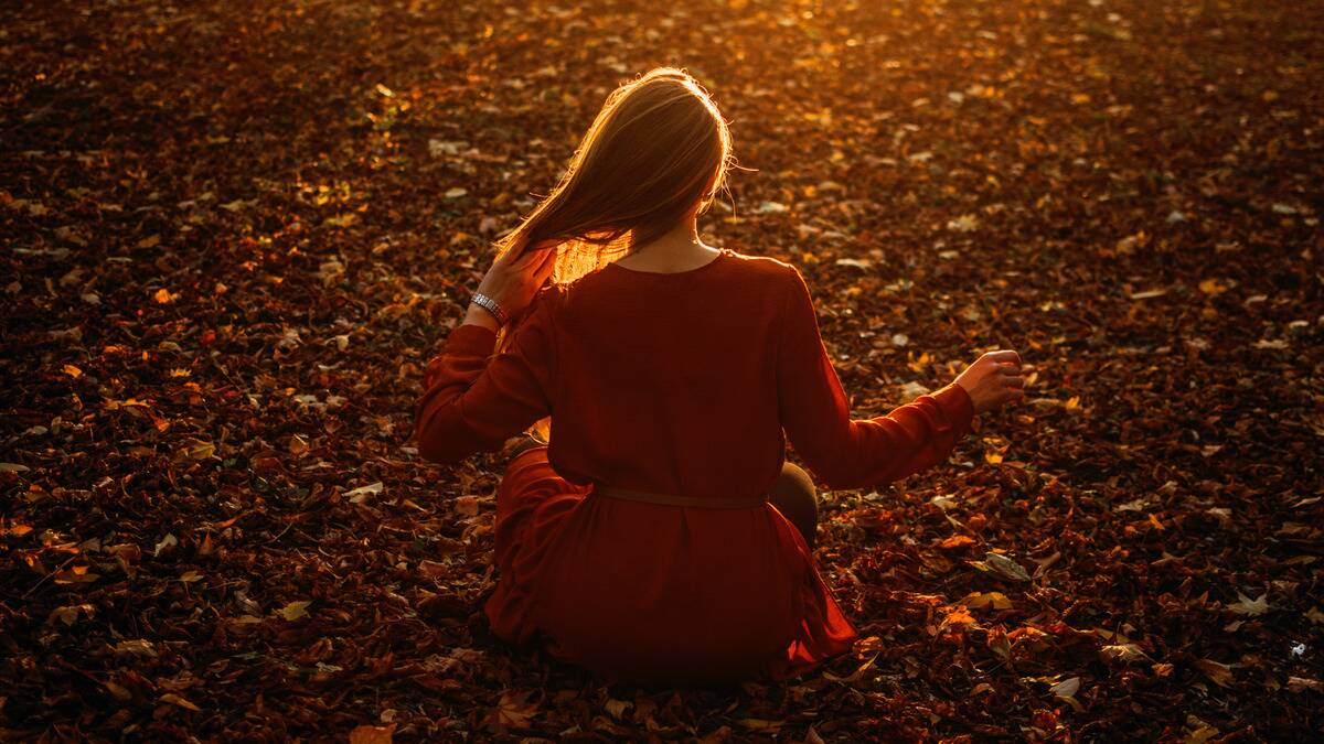 A woman sitting on the forest floor that's covered in autumn leaves, facing away from the camera, pulling her hair over her shoulder.