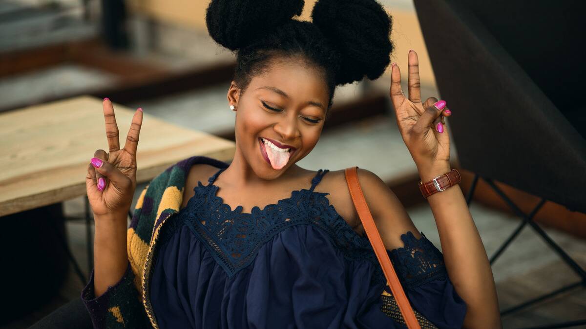 A young woman smiling with her eyes closed, sticking her tongue out, both hands up making a peace sign.