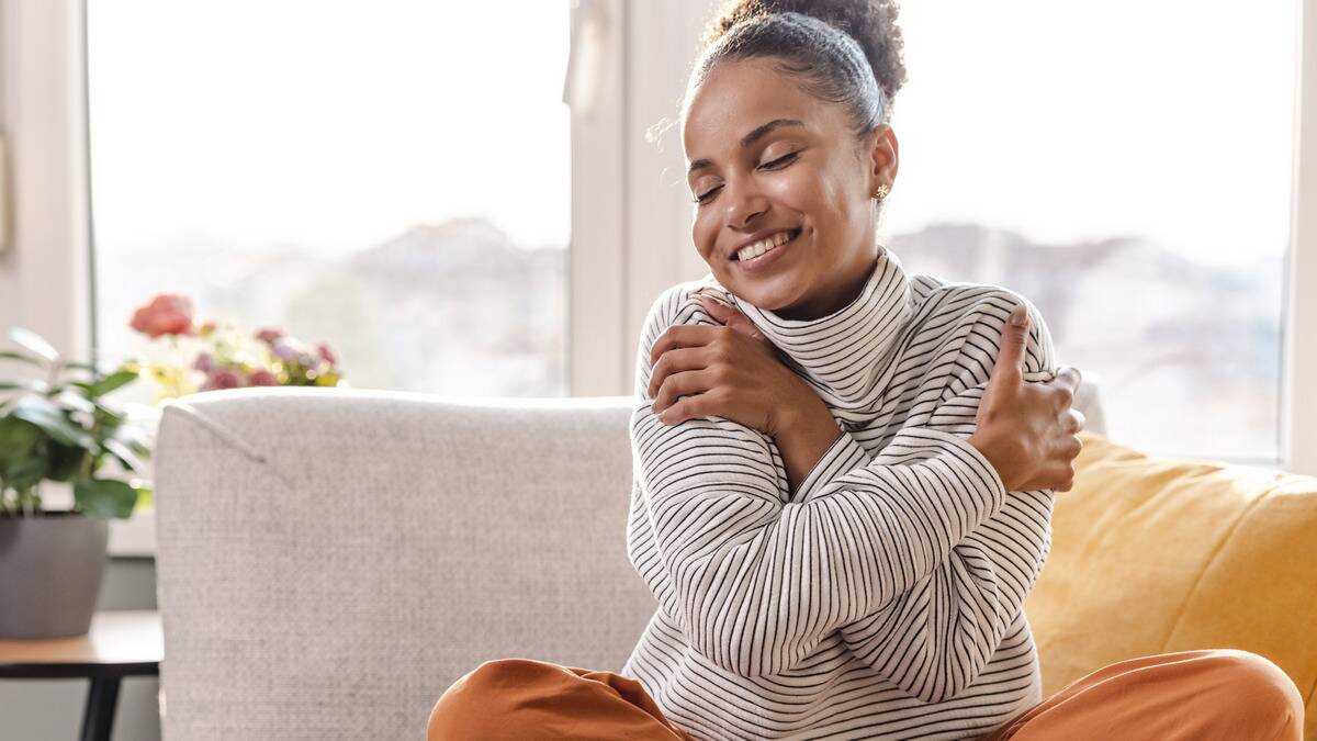 A woman sitting on her couch at home, smiling as she hugs herself.