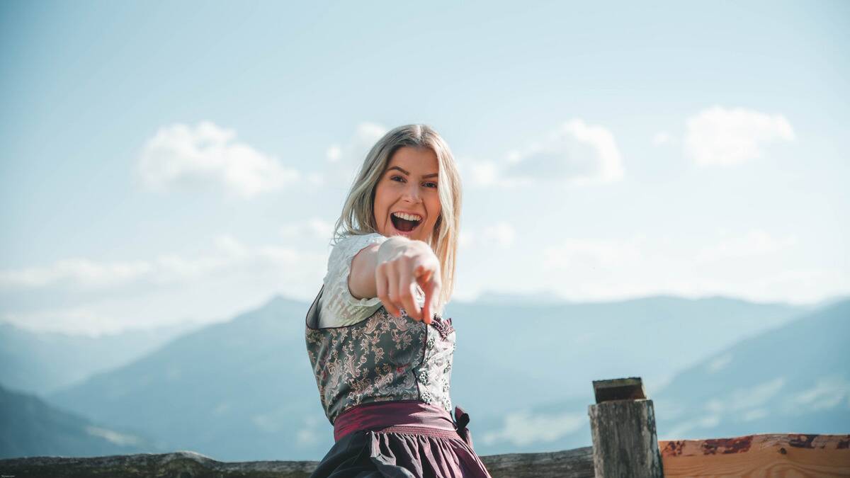 A woman standing outside under a clear blue sky, pointing at the camera that's angled a bit low, smiling brightly.