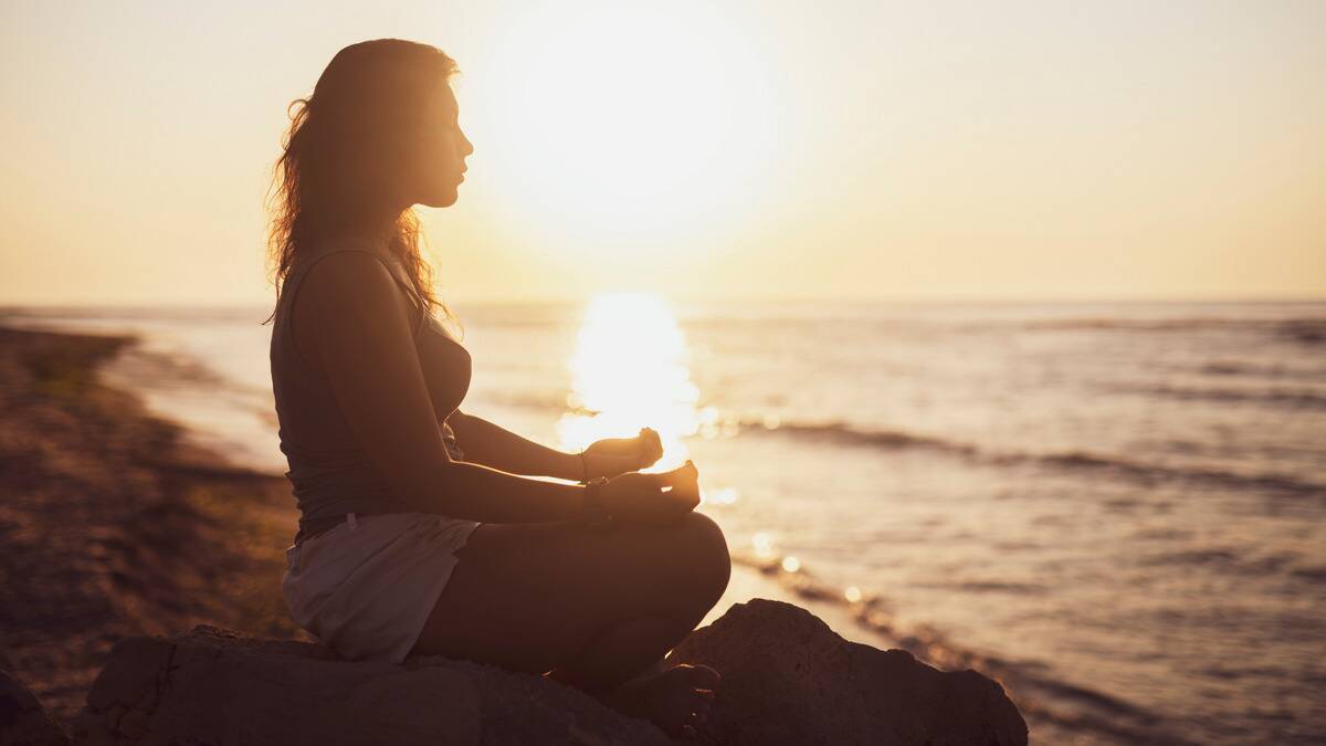 A woman sitting by the water meditating at sunset.