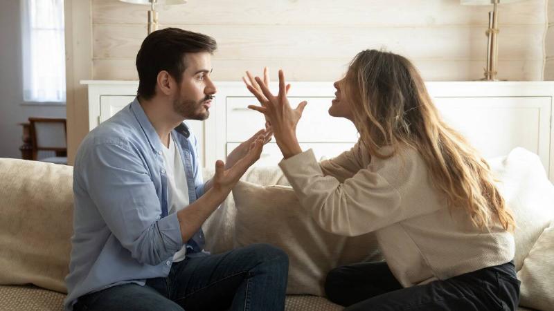 A couple sitting side by side on a couch but facing one another, yelling at each other, using their hands wildly as they speak.