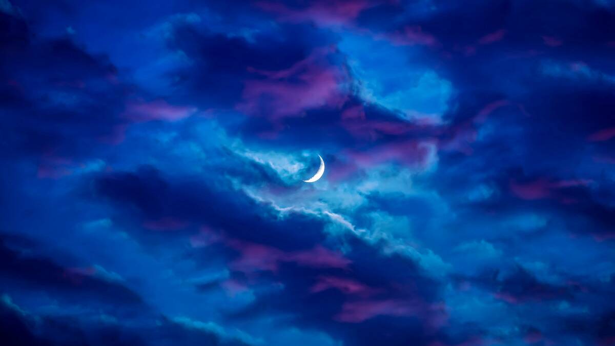 A small crescent moon seen amidst blue and purple colored clouds.