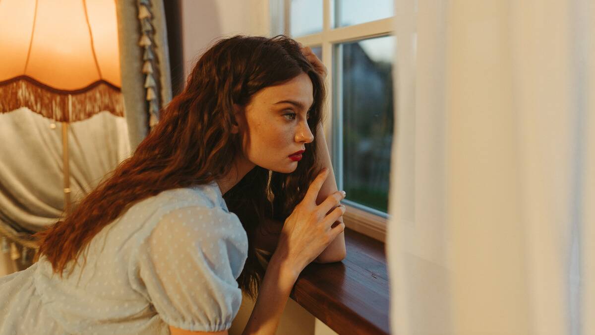 A woman leaning against a windowsill as she looks out the window, a hand in her hair.