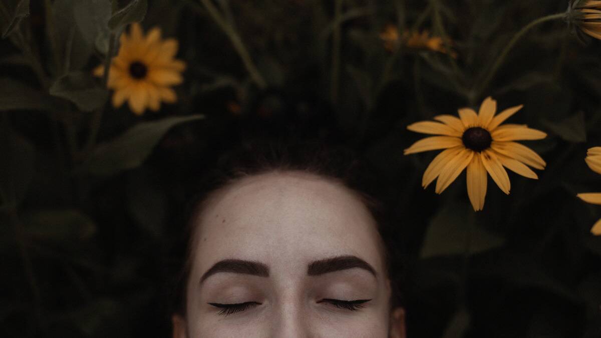 The top half of a woman's face seen laying in the grass, a few yellow flowers around her, her eyes closed.