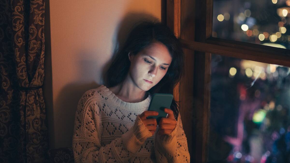 A woman sitting inside in the dark, her face illuminated by her phone screen that she's holding up.