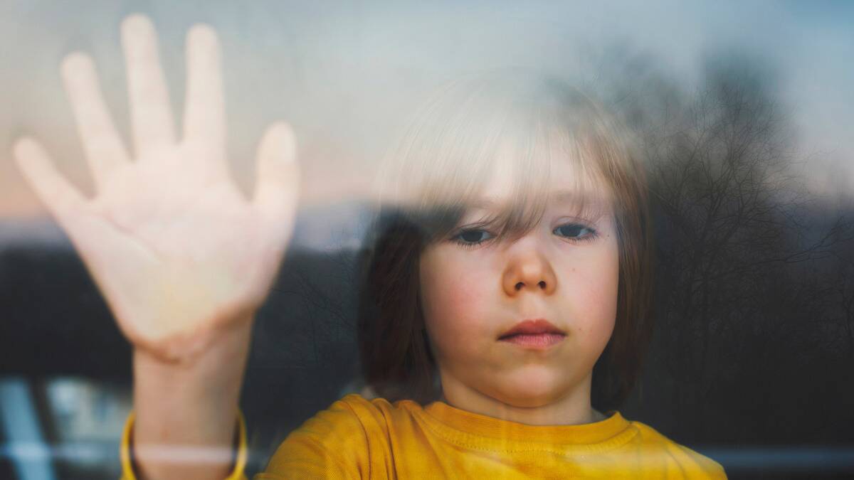 A young boy standing at a window, one hand pressed against the glass, looking sadly outside.