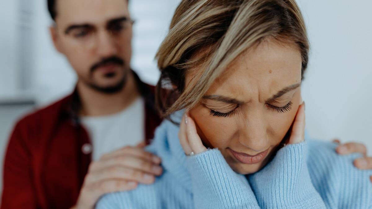 A woman looking stressed, her hands up covering the sides of her head, as her boyfriend stands behind her with his hands on her shoulders.