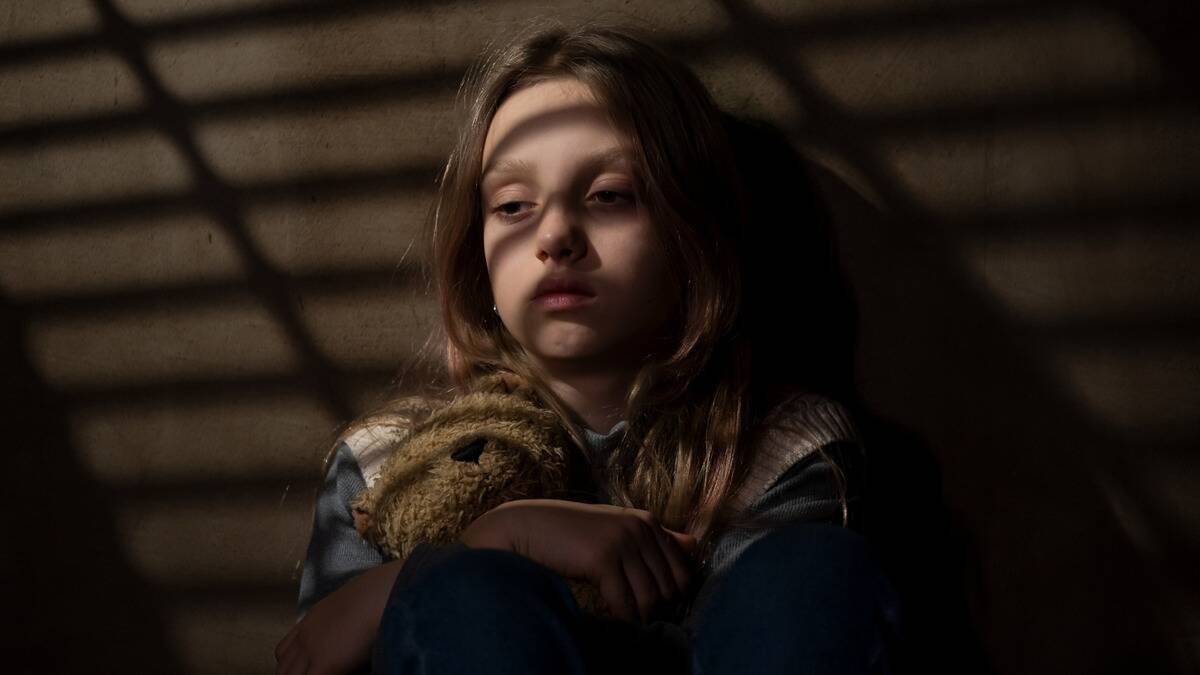 A young girl sitting against a wall in the dark, beams of light from a window shade across her face.