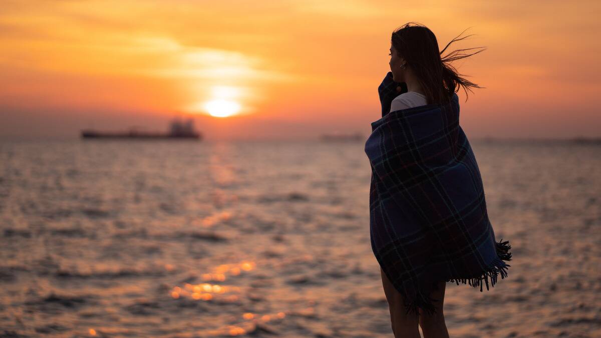 A woman standing outside by the lake at sunset, lookingo ut toward the horizon, wrapped in a blanket.