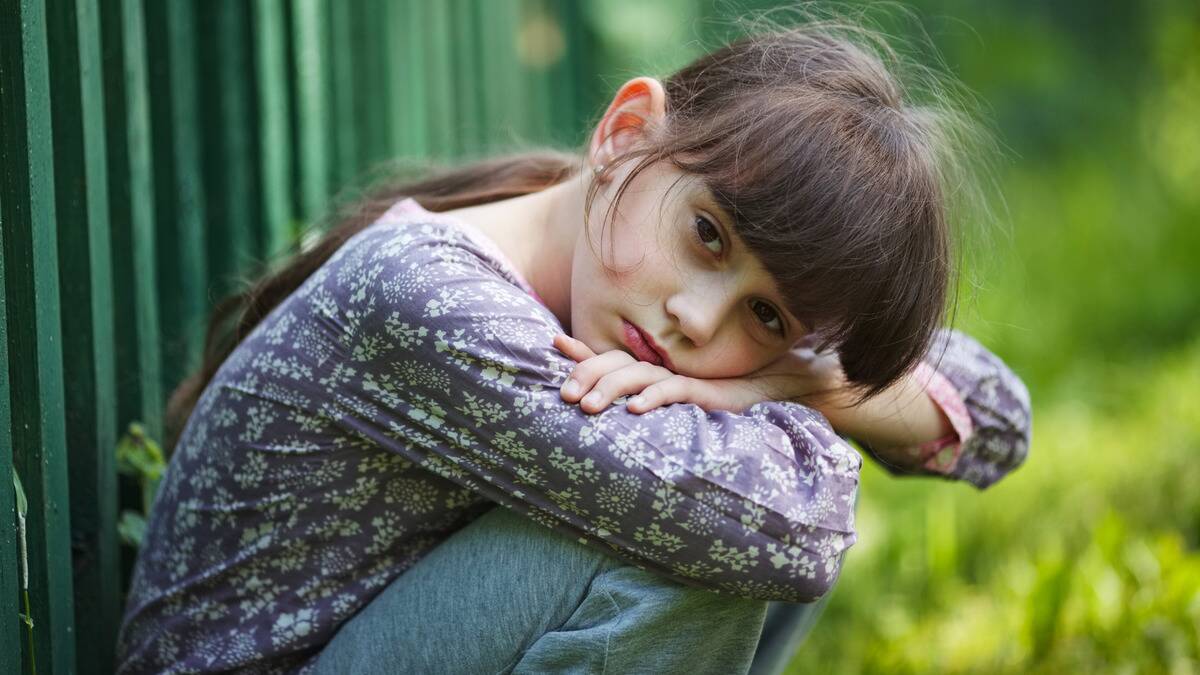 A young girl squatting on the ground, folding her arms over her knees, head resting on her arms, looking at the camera sadly.