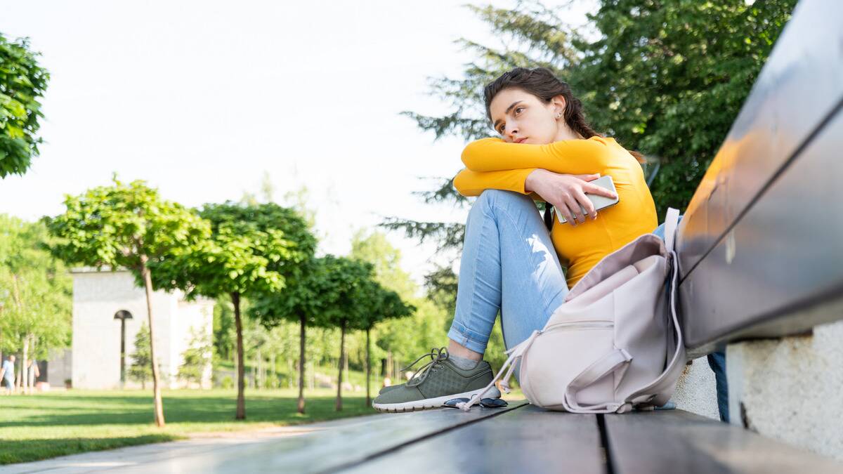 A woman sitting on a bench, knees drawn up, arms folded over them, face resting on her arms, looking off into the distance sadly.