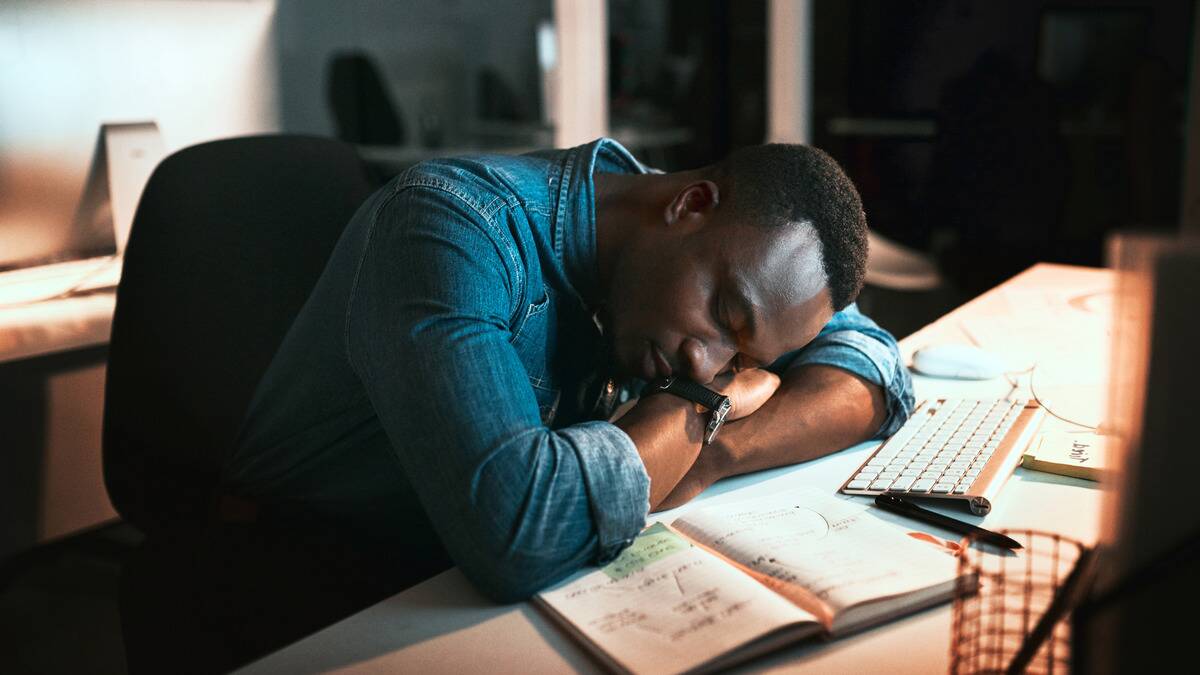 A man asleep at his desk, head on his arms, with papers and open notes around him.