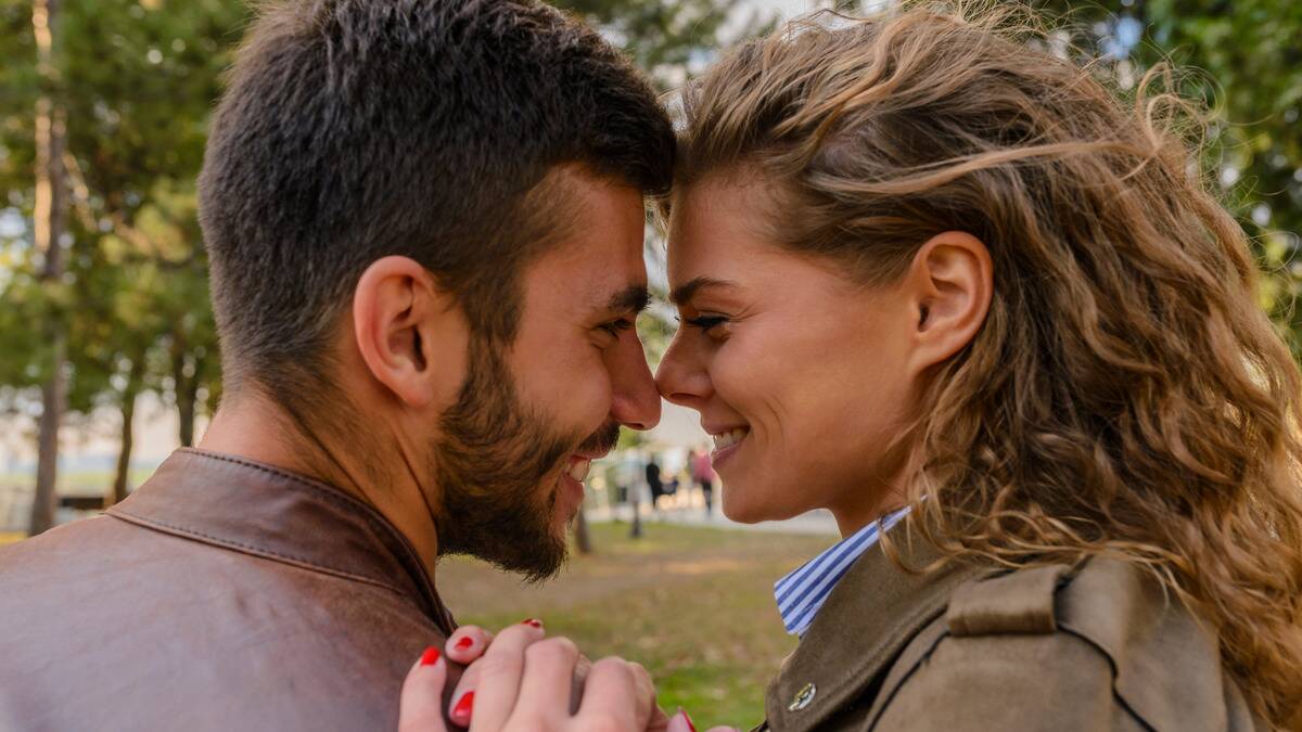 A close photo of a couple standing outside, looking at one another, their noses touching, both smiling.