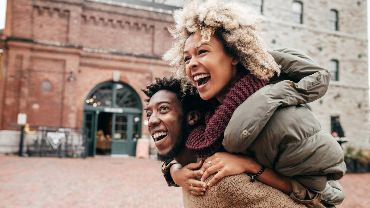 A couple both smiling as the man gives the woman a piggyback ride down the street.