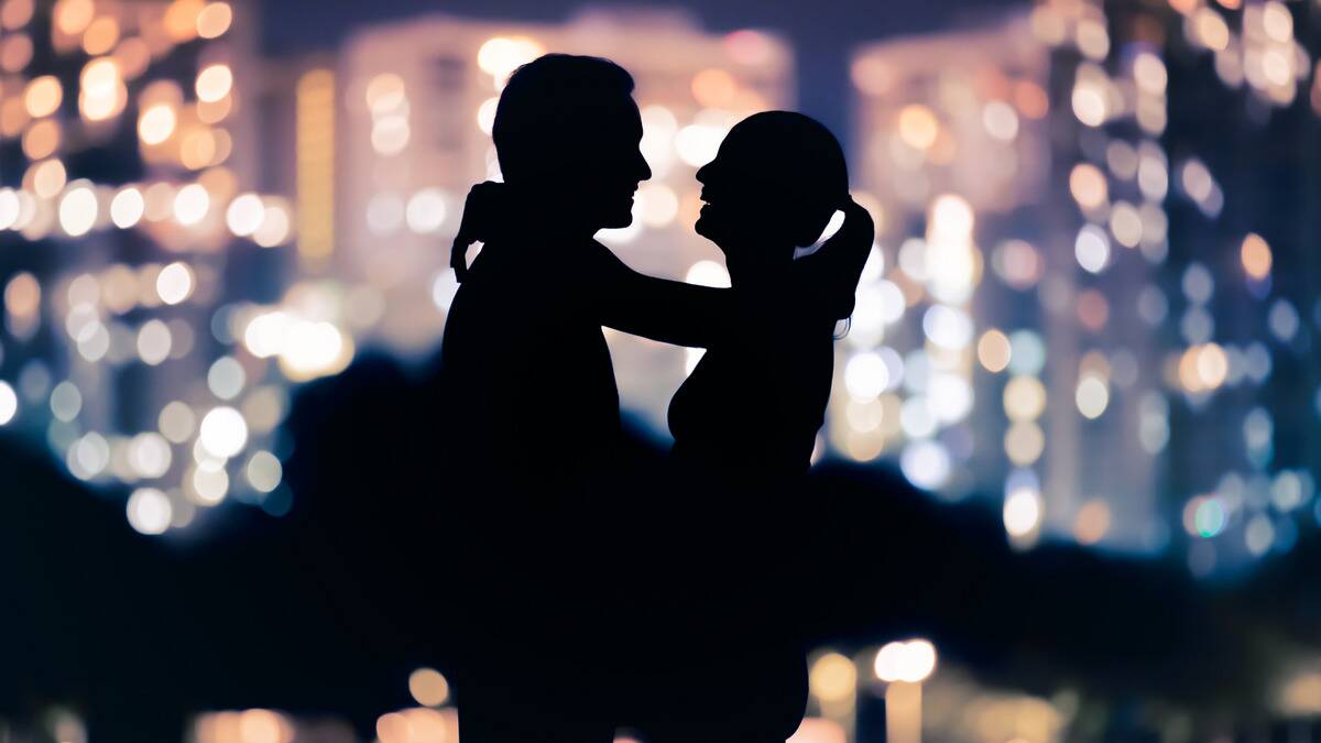 A silhouette of a couple standing in front of the city skyline, the woman with her arms around the man's neck.