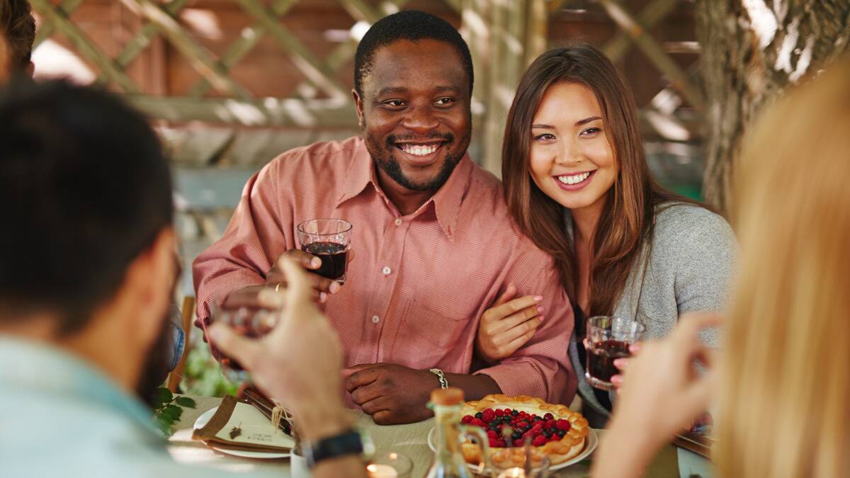 A couple sitting together at a dinner, each holding a glass, the woman's arm hooked beneath the man's.