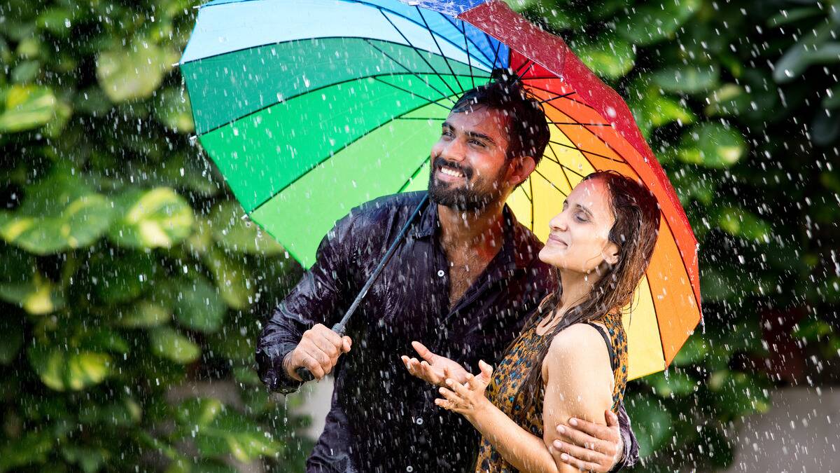 A couple standing outside in the rain under a large rainbow umbrella, though they're both still wet. Both are smiling.