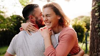 A couple hugging outside, both smiling, the woman with her eyes closed as they both hang on tightly.