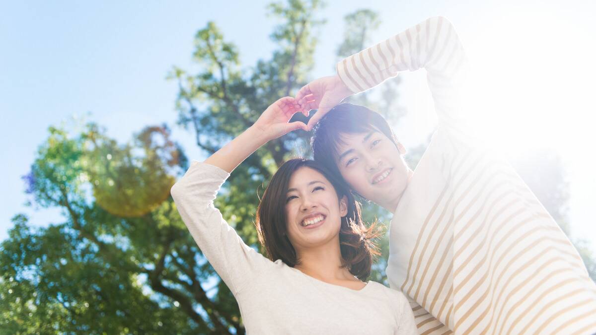 A couple standing outside, making a heart with their hands together, the sun shining down on them.