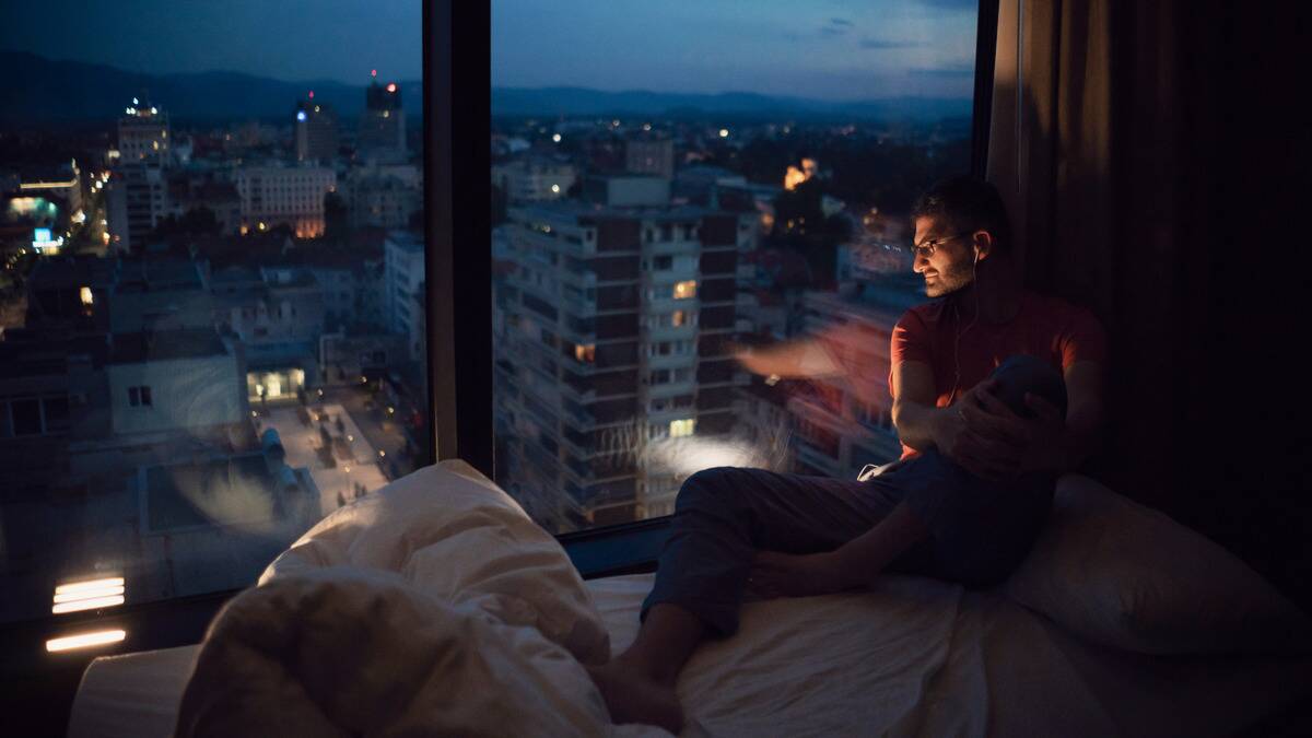 A man sitting by the large window of his high-rise apartment, one knee pulled up, looking out at the horizon.