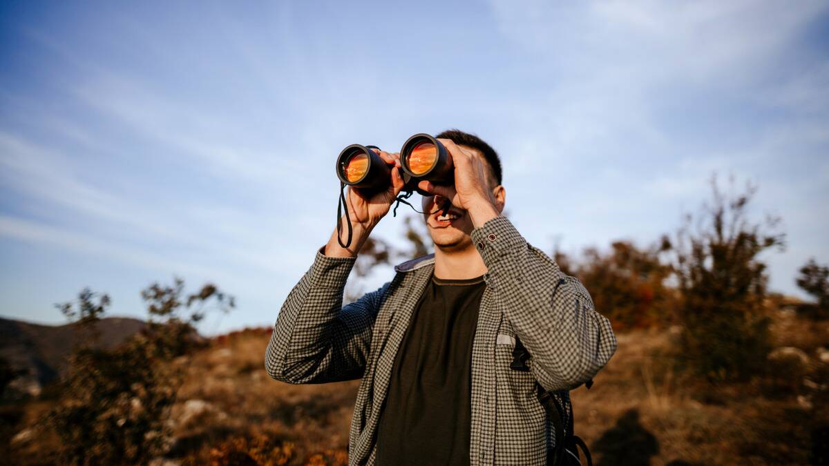 A man standing on a hill looking ahead with large binoculars.