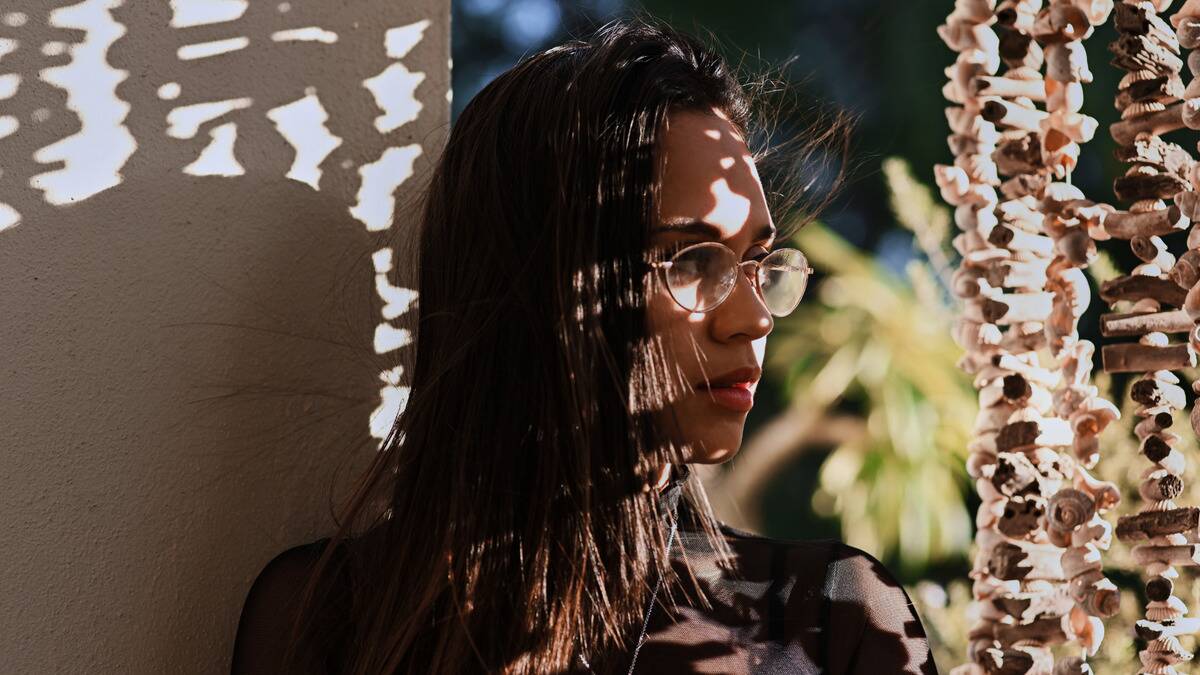 A close photo of a woman sitting outside, mostly in shadow, but with specks of light shining on her face as she looks off to the side.