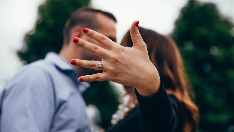 A couple kissing, but their faces are being hidden by the woman's hand in the foreground showing off her new engagement ring.