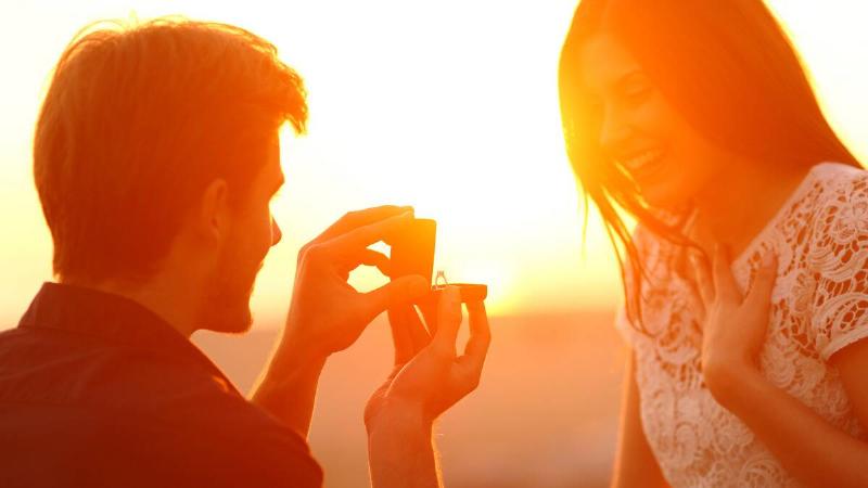 A close shot of a man on one knee proposing to his girlfriend, who's smiling with a hand on her chest, as the sun sets behind them.