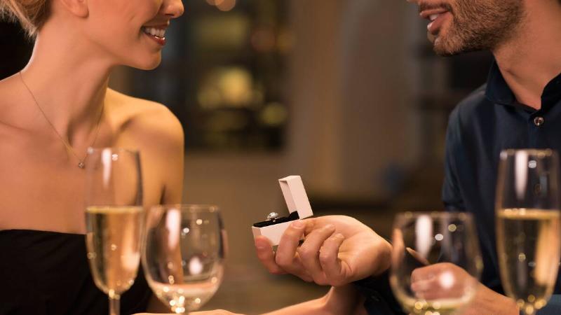 A man proposing to his girlfriend at a dinner table, holding up a ring box.