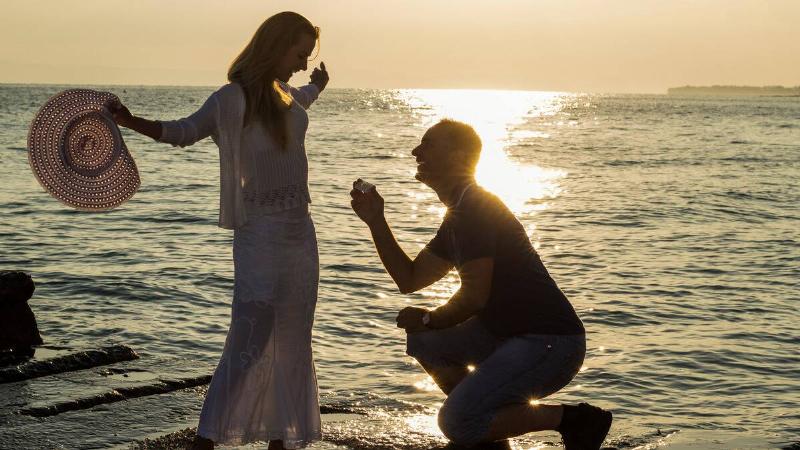 A man on one knee proposing to his girlfriend while they stand by the water, the woman standing with her arms out.