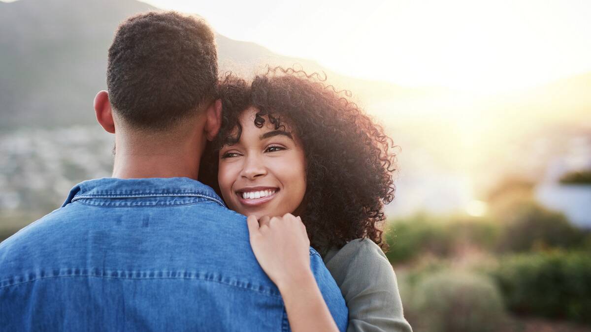 A couple hugging, we can see the man's back and the woman's face on his shoulder, smiling.