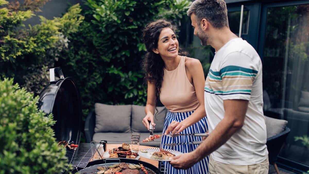 A couple standing by a grill together, plating food they've made, smiling at each other as they do.