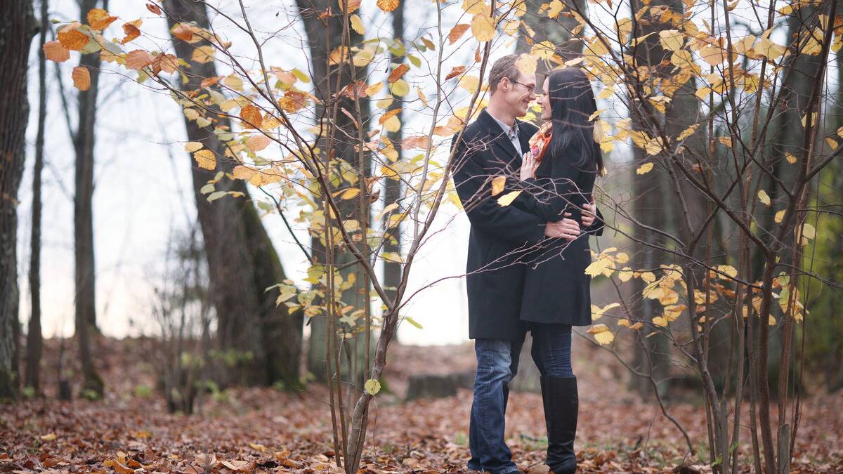 A couple standing outside amid autumn leaves, holding each other close, smiling at one another.