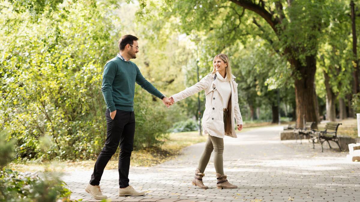 A couple holding hands walking outside, the woman leading a little and looking over her shoulder at the man, the two smiling at each other.