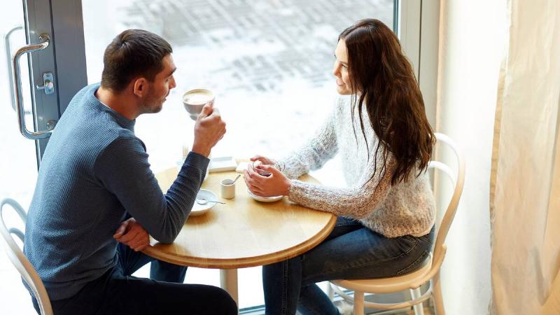 A couple out for coffee, sitting opposite each other at a small table, the woman chatting as the man raises his cup to drink.