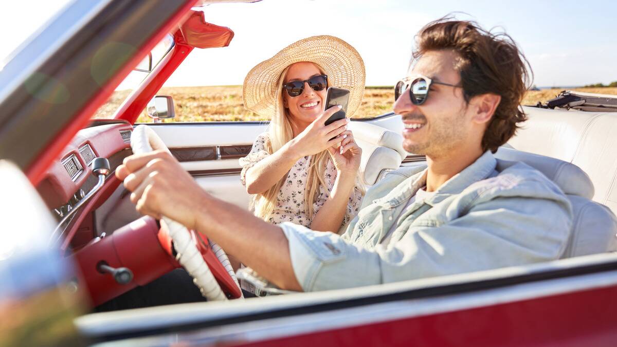 A couple on a road trip in a convertible car, the woman taking a photo of the man, who's driving, with her phone.