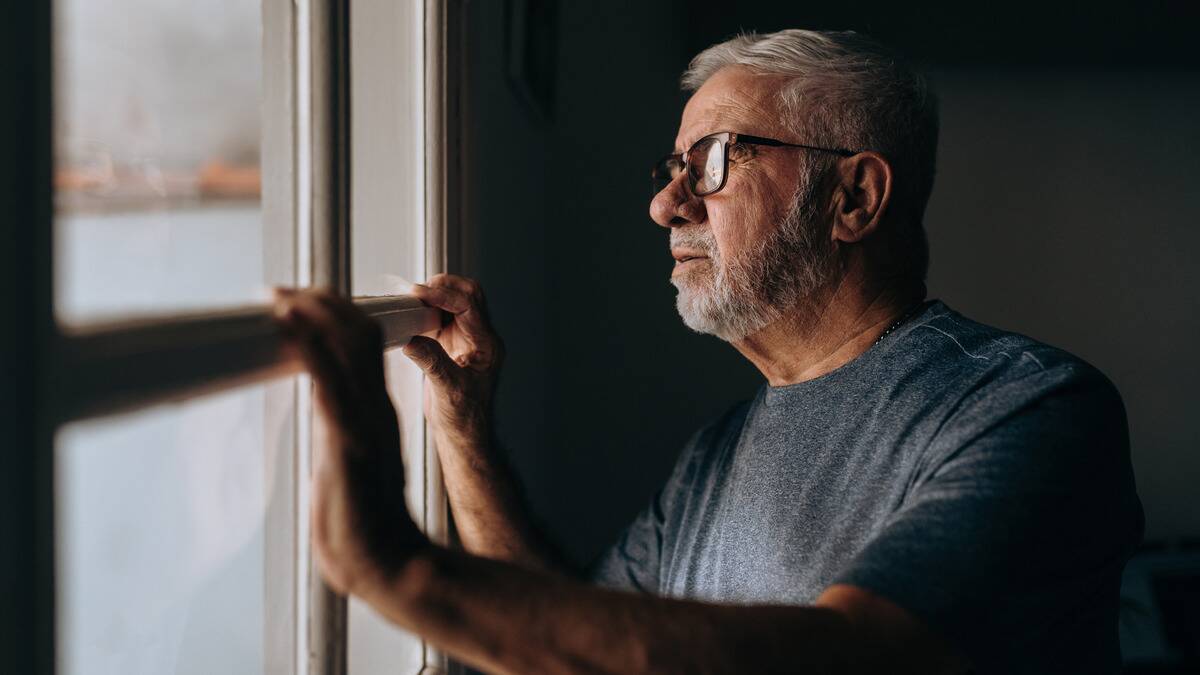 A man looking out the window of his home, both hands resting on the windowframe.