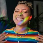 A young person smiling brightly as beams of rainbow light shine upon their face.