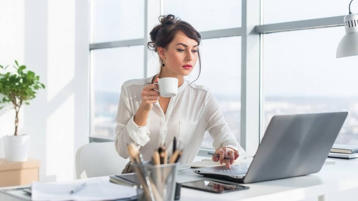 A put-together business woman working away at her desk, holding up a mug as she does.