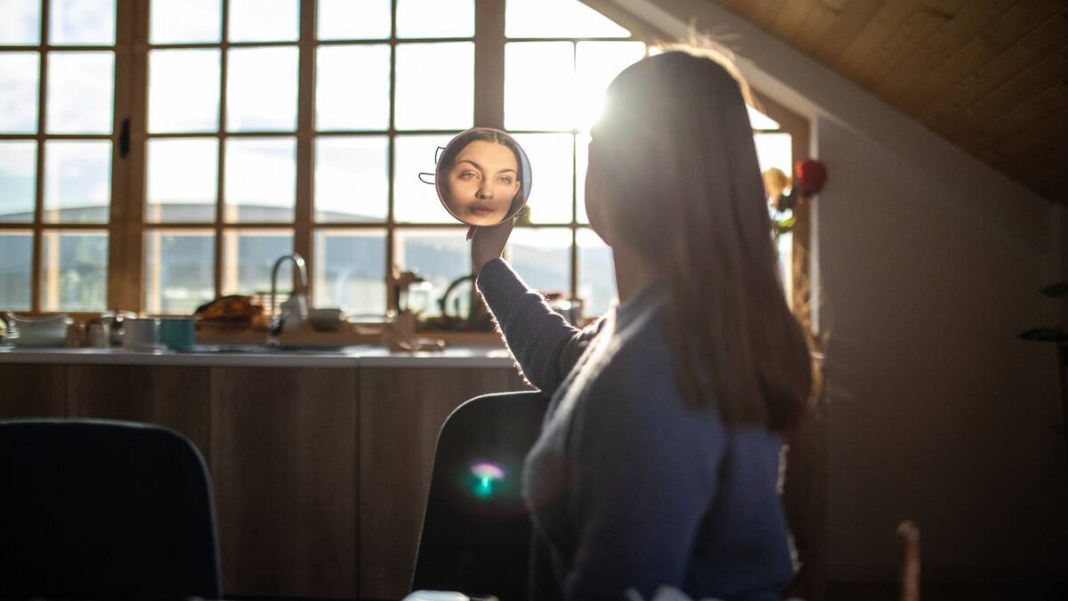 A woman, facing away from the camera toward a large window, holding up a round mirror that we can see her face in the reflection of.